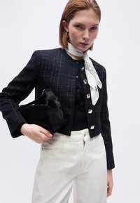 Γυναίκα με μαύρο υφασμάτινο κοντό σακάκι με χρυσά κουμπιά, λευκό παντελόνι, λευκό φουλάρι με σχέδια γύρω από το λαιμό, κρατώντας μαύρη τσάντα από γούνα.