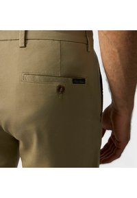 Pantaloni khaki con una texture liscia, dotati di una tasca posteriore con chiusura a bottone e un piccolo etichetta con il marchio. La vista laterale mostra la vestibilità e la forma.