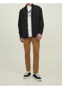 Svart skjorta med knappar över en vit t-shirt med grafik, khakibyxor och svarta sneakers med vita sulor. Enkelt och avslappnat design.