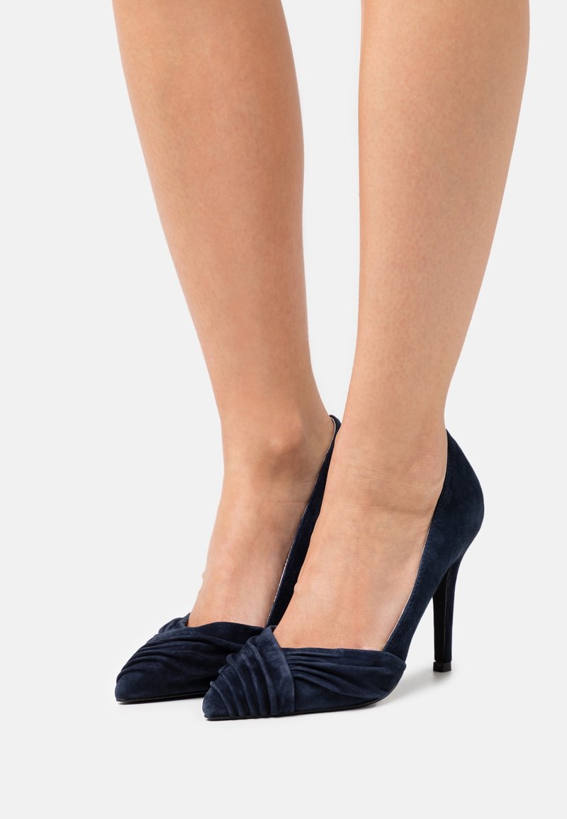 Paire d'escarpins en suède bleu marine avec détails pliés sur les bouts pointus, portés pieds nus sur un fond uni.