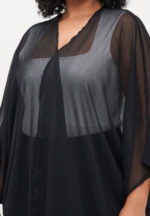 Vrouw draagt een zwarte doorschijnende overlay met wijde mouwen over een zilverkleurige gestructureerde top, met gelaagde kleding en een contrast in stof.