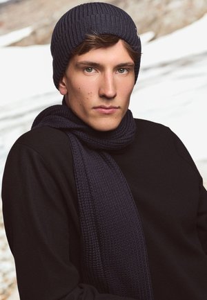 Navy knit beanie and scarf paired with a black long-sleeve top. The beanie has a ribbed texture; the scarf features a woven pattern.