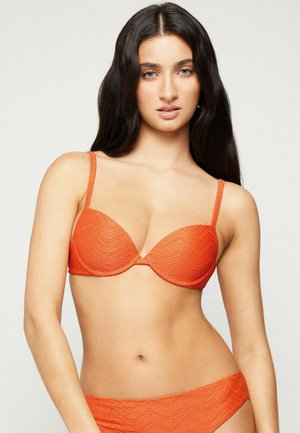 Woman with long dark hair wearing orange textured bikini top and bottom, standing against a plain light background.