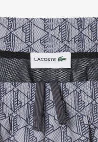 Shorts à taille élastique avec motif géométrique gris, doublure en mesh noir, cordons de serrage gris, et étiquette Lacoste blanche avec logo crocodile vert.