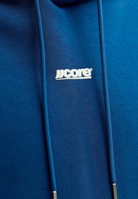 Close-up van blauwe hoodie-stof met blauwe trekkoorden en een klein wit "JJcore"-logo in het midden van de borst.