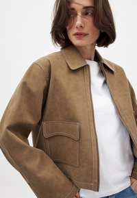Giacca in suede marrone con collo, zip frontale, tasche sul petto doppie e dettagli di cucitura bianca. Indossata su una camicia bianca.