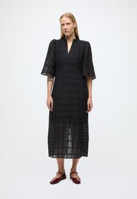 Juoda, iki kelių siekianti suknelė su V formos iškirpte ir pūstomis rankovėmis. Tekstūruota medžiaga su tinklelio raštu. Derinama su raudonais lygiais bateliais.