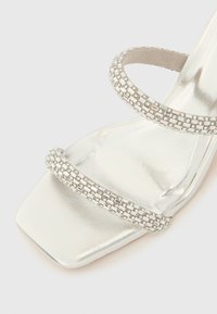 Ασημί σανδάλι με τακούνι και τετράγωνη μύτη, με δύο λουριά διακοσμημένα με μικρές διαφανείς στρας και μεταλλική λεία επιφάνεια.