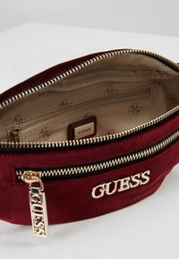 Bolsita de terciopelo color burdeos con herrajes dorados, que incluye un bolsillo exterior con cremallera, compartimentos interiores y una etiqueta con el logo. Forrada con tela beige estampada.