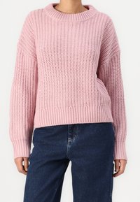 Różowy sweter dzianinowy z wysokim kołnierzem, oversize'owy fason oraz ribbowymi mankietami i dołem; teksturowany materiał o luźnym splocie.