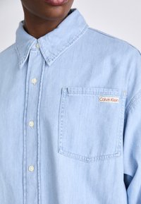 Lyseblå denimskjorte med knaplukning, krave og en venstre brystlomme med et rødt og hvidt Calvin Klein-label.