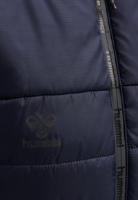 Padded granatowa kurtka o strukturze kratki, z czarnym zamkiem błyskawicznym i logo. Materiał wydaje się gładki i lekki.