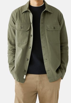 Man draagt een olijfgroen jack met knopen over een zwarte trui en beige ribfluwelen broek, met zijn handen in de zakken.