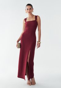 Robe ajustée bordeaux avec un décolleté carré, en tissu texturé, fente sur le côté et bretelles larges ; accessorisée avec des boucles d'oreilles en or et un petit sac à main.
