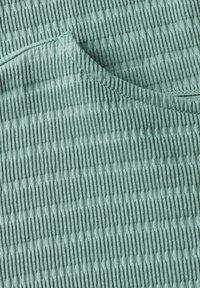 Texturovaná zelená látka s vertikálními pruhy a hladkými oblastmi, ukazující měkkou pletenou kvalitu. Viditelný kulatý výstřih.