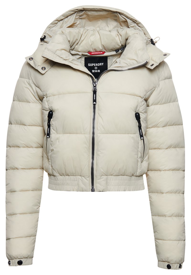 Giacca a vento corta in beige chiaro con cappuccio, chiusura con zip e due tasche laterali con zip. Presenta un bordo a coste e polsini con bottoni a pressione.