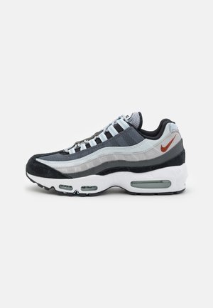 Vista lateral de una zapatilla Nike Air Max 95 en tonos gris, negro y blanco, con cojines de aire visibles y el logo de Nike en color naranja.