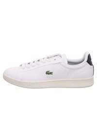 Baskets blanches basses avec lacets blancs, logo crocodile vert sur le côté et languette de talon noire, présentées sur un fond blanc.