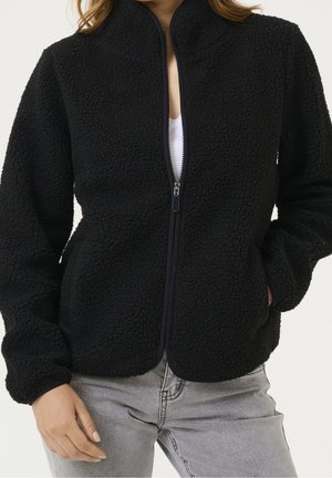 Black textured fleece jacket with zipper front, worn over white top and paired with light gray jeans, person’s hands relaxed by sides.