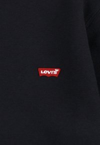 Tecido preto com uma textura suave, apresentando uma pequena etiqueta com o logótipo vermelho da Levi's numa forma clássica do lado esquerdo.