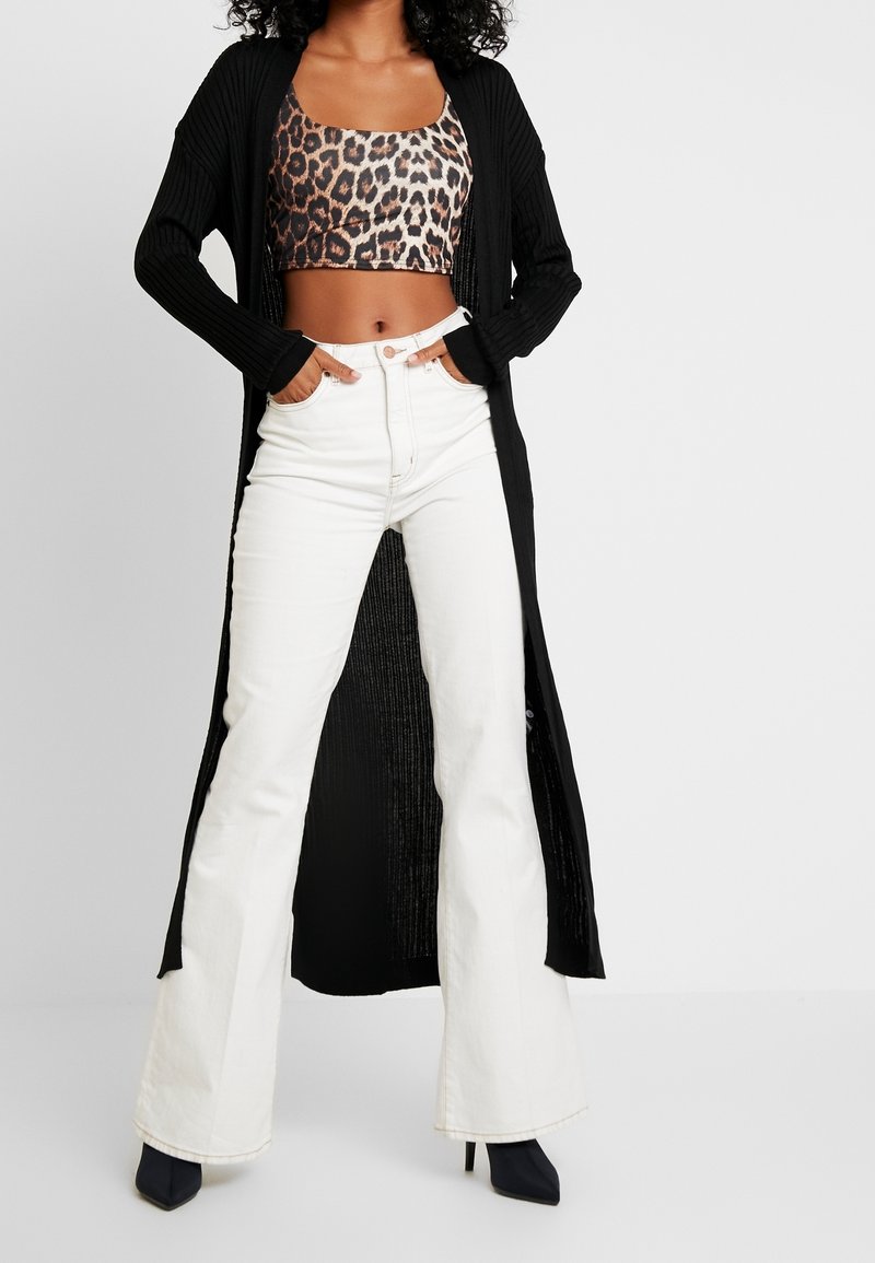 Lång svart ribbad cardigan över en leopardmönstrad crop top, ihop med vita utsvängda jeans och svarta klackskor.