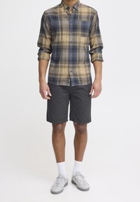 Chemise en flanelle à carreaux jaunes, noirs et bleus, associée à un short noir. Le mannequin porte des chaussettes blanches et des baskets grises. Manches courtes retroussées.