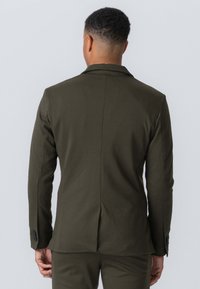 Olivengrøn skræddersyet blazer med strukturerede skuldre, enkelt vent bagpå og knapper ved ærmerne. Glat tekstur med et sofistikeret design.