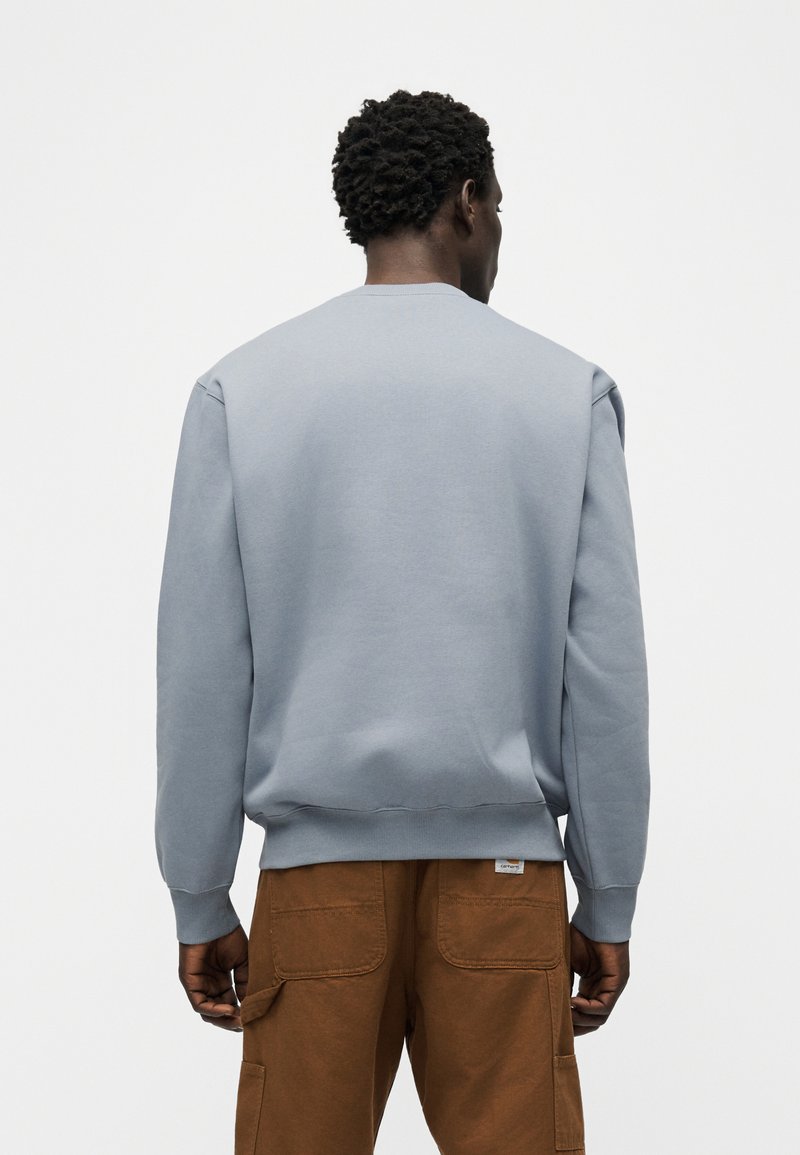 Mann mit kurzen lockigen Haaren, der einen hellgrauen Pullover und braune Cargohosen trägt, steht mit dem Rücken zu einer einfarbigen Wand.