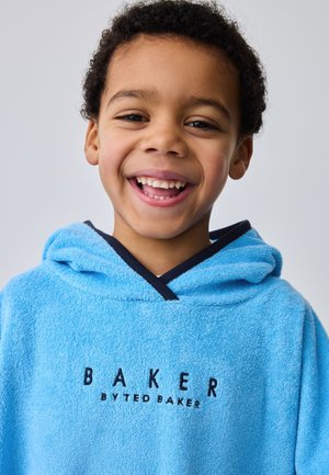 Uśmiechnięty chłopiec o kręconych włosach, ubraną jasnoniebieski ręcznik z kapturem z napisem „Baker by Ted Baker” z przodu na jednolitym tle.