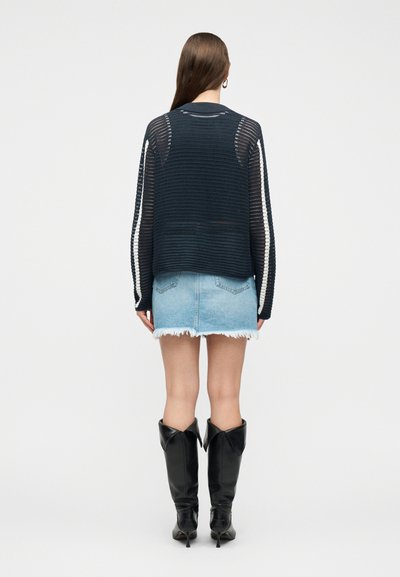 Γυναίκα που φοράει μαύρο μακρυμάνικο ανοιχτό πλεκτό τοπ, ανοιχτό μπλε μίνι φθαρμένη τζιν φούστα και μαύρες μπότες έως το γόνατο με μυτερή μύτη, γυρισμένη με την πλάτη.
