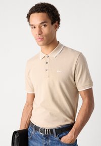 Beige polotrøye med hvit krage, tre knapper, og en liten logo på brystet. Båret med mørke denimjeans og et svart belte.