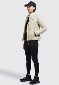 Jachetă crem căptușită, cu un design în valuri, purtată peste un top negru și colanți. Adidași negri transparenti și o șapcă neagră completează look-ul.
