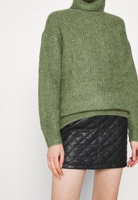 Pull en maille verte à col roulé avec un motif texturé, associé à une mini-jupe en cuir matelassé noire, mettant en valeur une silhouette ajustée.
