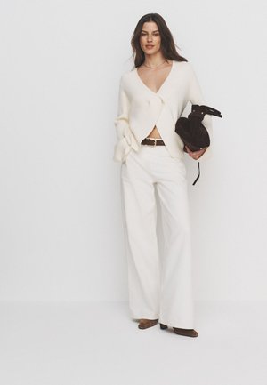Femme portant un cardigan crème et un pantalon blanc à jambes larges, tenant un sac à main marron foncé, debout devant un fond blanc uni.