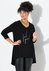 Schwarzes V-Ausschnitt-Tunika mit Dreiviertelärmeln, versehen mit silbernem Strassschriftzug "feel good". Hergestellt aus weichem Material, mit einem asymmetrischen Saum.