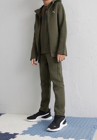 Olivengrøn træningsdragt med lynlåshætte og matchende bukser. Har en logo på brystet, ribstrikkede detaljer og sorte sportssko.