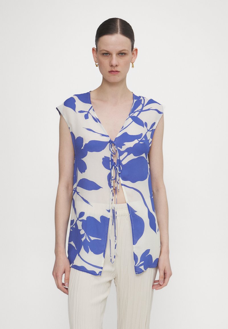 Blauw bloemenpatroon-top met een V-hals en strik aan de voorkant. Korte mouwen, lichtgewicht stof en een open ontwerp. Gecombineerd met lichte broek.