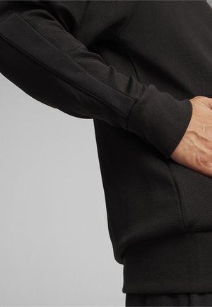 Crna dukserica s teksturiranim materijalom, rebrastim rukavima i bočnim džepom. Sadrži kontrastne šavove duž rukava.