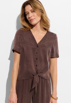 Kobieta o blond włosach do ramion, ubrana w brązową krótko rękawową bluzkę zapinaną na guziki, związaną w talii, oraz pasujące spodnie.