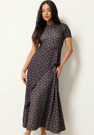 Frau trägt ein schwarz-graues Polka-Dot-Kurzarmkleid mit asymmetrischem, geschichtetem Rock vor hellem, einfarbigem Hintergrund.