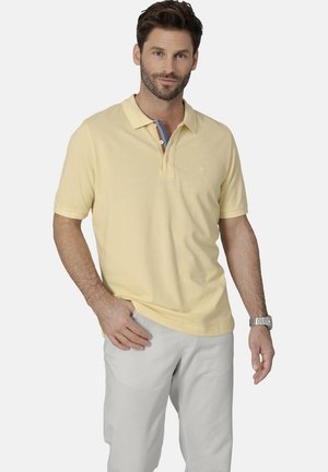 Gelbes Poloshirt mit Kragen und drei Knöpfen, mit einem mehrfarbigen Streifendetail unter dem Kragen. Getragen mit hellgrauen Hosen.