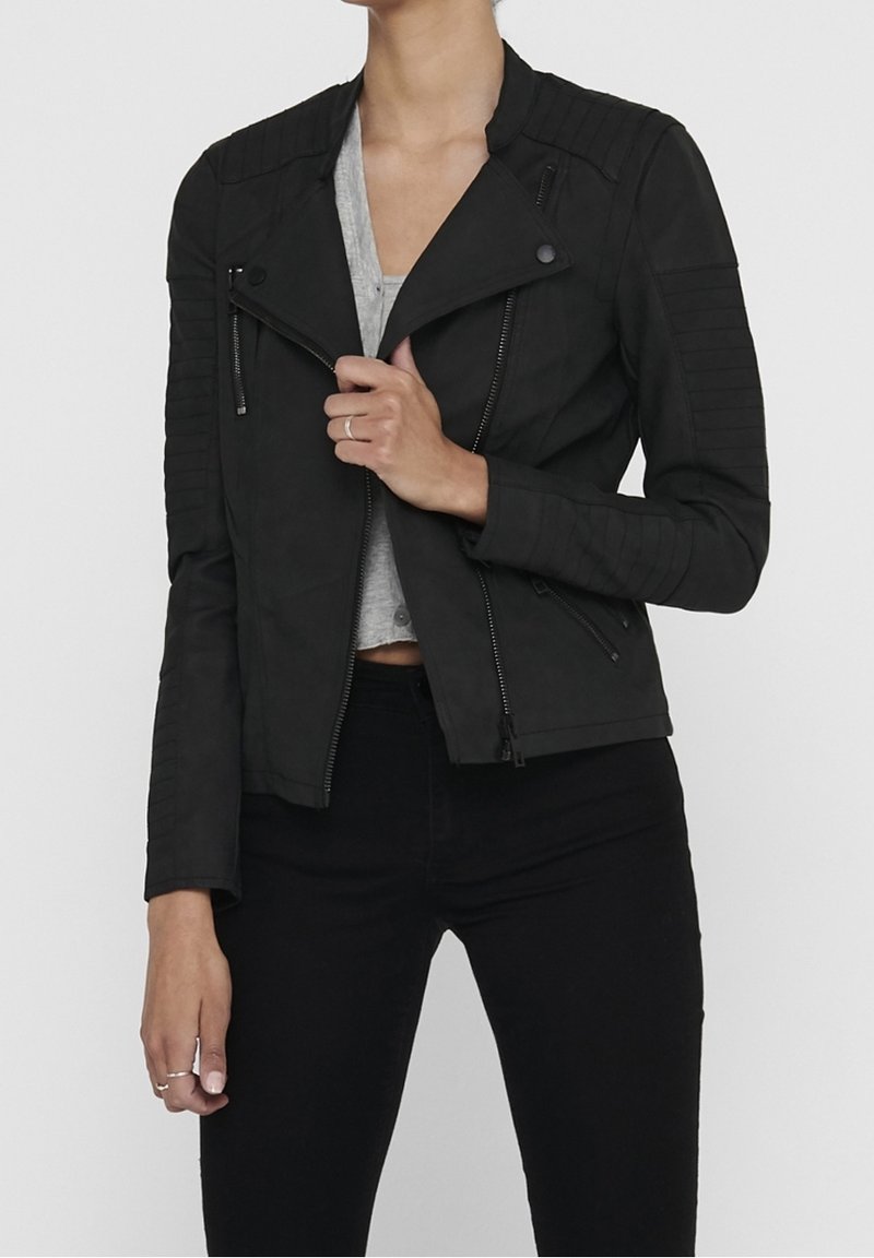 Veste de moto en simili cuir noir avec des fermetures éclair, des accents côtelés sur les épaules et une coupe ajustée. Portée sur un haut gris et un pantalon noir.