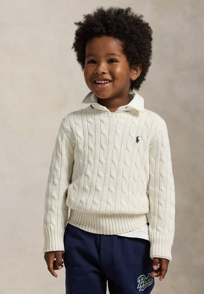 Smilende ung dreng med krøllet hår iført en hvid kabelstrikket sweater over en skjorte med krave og marineblå Polo-bukser.