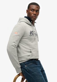 Grijze hoodie met een voorzak, trekkoordkap en marineblauwe logo. Gecombineerd met een spijkerbroek, die een ontspannen pasvorm en katoen materiaal laat zien.
