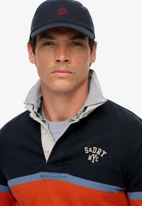 Marineblauw en oranje gestreepte rugbyshirt met een kraaghalslijn, voorzien van knoopdetails en geborduurd "S&DNYC" logo. Pet gedragen op het hoofd.