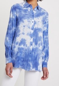 Chemise à manches longues bleue tie-dye avec une fermeture boutonnée à l'avant, présentant un ourlet incurvé, un col et des poignets froncés ; fabriquée en matériau léger.