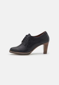 Zapato de cuero negro con punta redondeada, parte delantera con cordones y tacón medio apilado. Textura suave con mínimo detalle de costura.