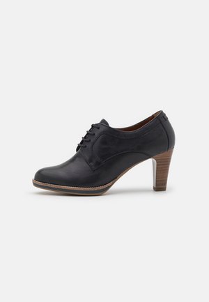 Zapato de cuero negro con punta redondeada, parte delantera con cordones y tacón medio apilado. Textura suave con mínimo detalle de costura.
