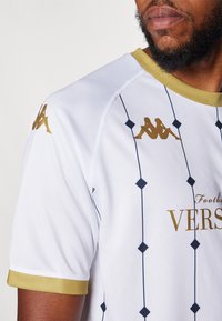 Vit fotbollströja med guldaccenter och en strukturerad yta. Den har en guldkrage, sidografik och diamantmönster i marinblått.
