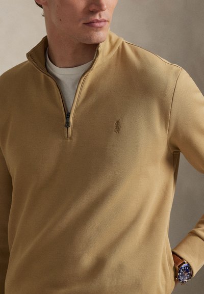 Άνδρας που φοράει μπεζ πουλόβερ με φερμουάρ μέχρι τη μέση πάνω από λευκό πουκάμισο, δείχνοντας το κεντημένο λογότυπο στο στήθος και το ρολόι με μπλε καντράν στον καρπό.